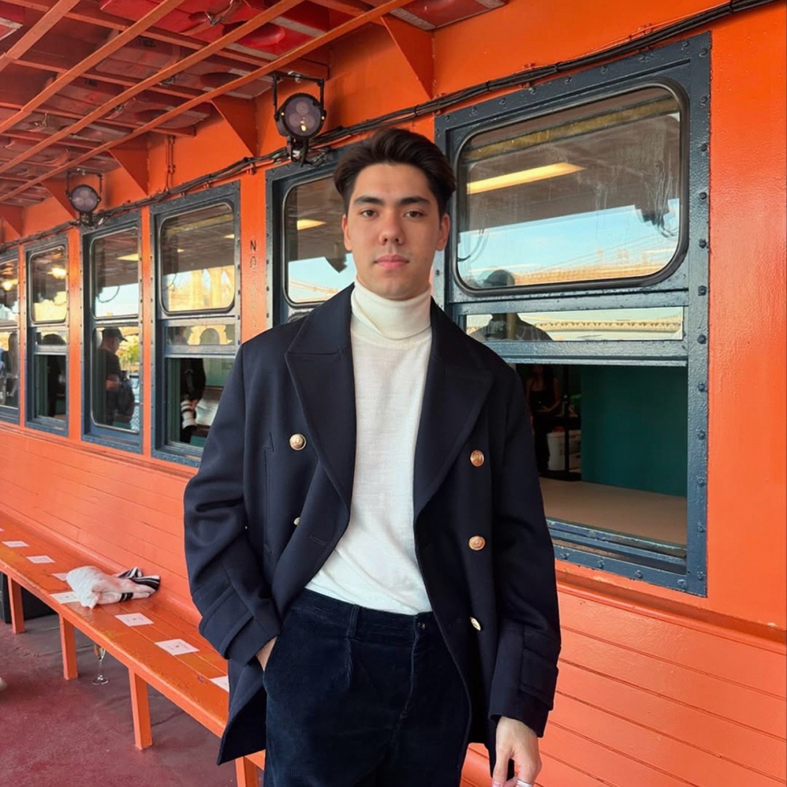 Matthew Stevens in a navy double-breasted coat and white roll-neck in front of a bright orange wall