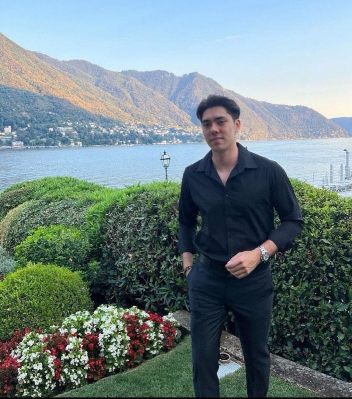 Matthew Stevens dressed in black above a lake and mountains, with flower beds in front