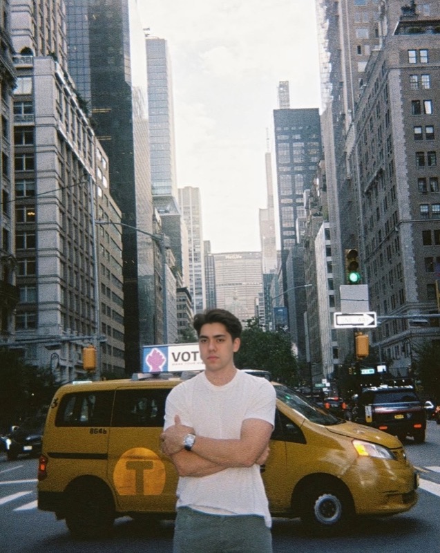 Matthew Stevens in a white T-shirt, arms crossed, on a New York avenue beside a yellow cab