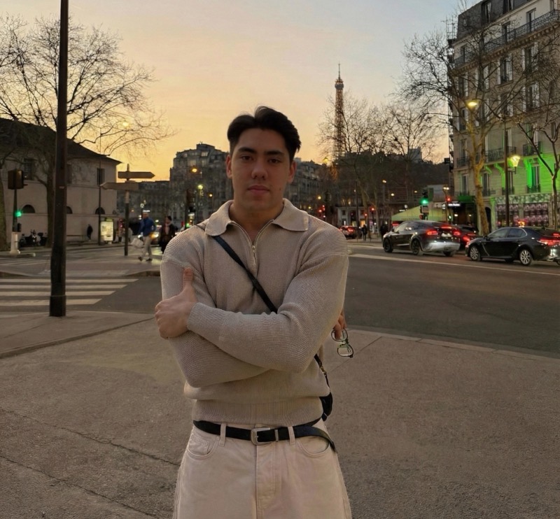 Matthew Stevens in a knit polo, arms crossed, on a Paris street at sunset with the Eiffel Tower behind