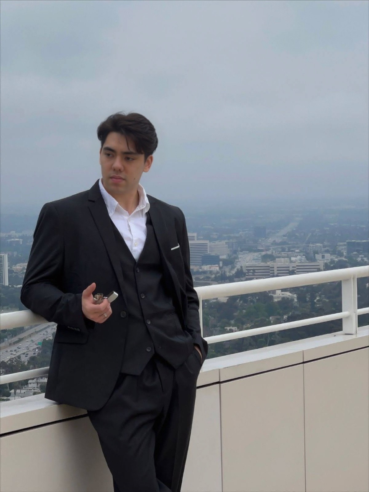 Matthew Stevens in a black three-piece suit leaning on a rooftop railing above a hazy city