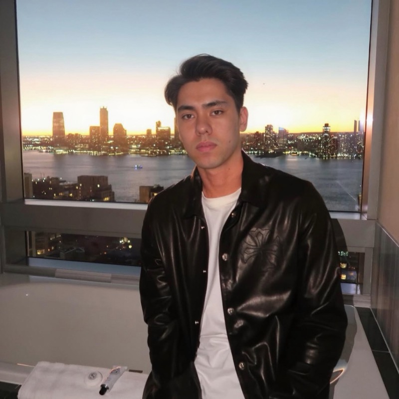 Matthew Stevens in a black leather jacket, with a city skyline across the water at sunset